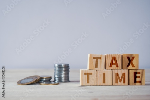 Wooden blocks with text sign TAX TIME placed on desk with stack of coins. Selective focus on the text, copy space.