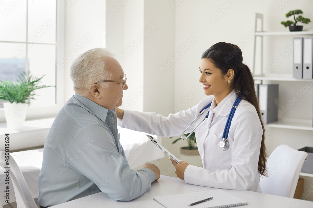 Fototapeta premium Friendly smiling young female doctor patting on shoulder of senior patient sitting at desk telling good analysis test and effective treatment results. Medicine and health insurance for elderly people