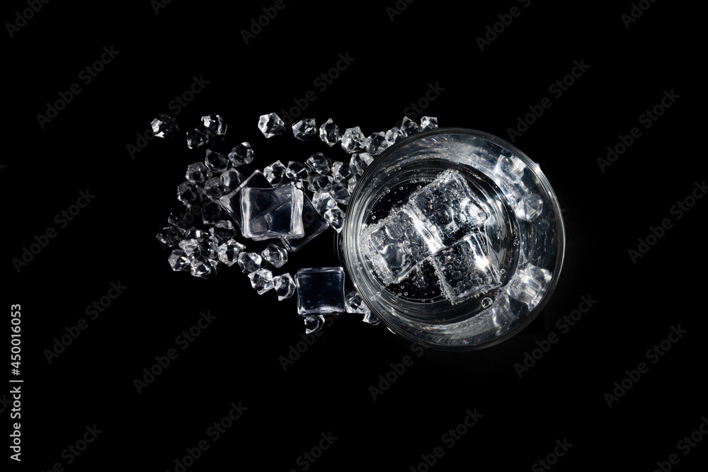 A transparent glass beaker with a colorless liquid ice cubes floating in it and scattered on the black surface on which the glass stands, view from above A transparent glass beaker with a colorless liquid ice cubes floating in it and scattered on the black surface on which the glass stands, view from above