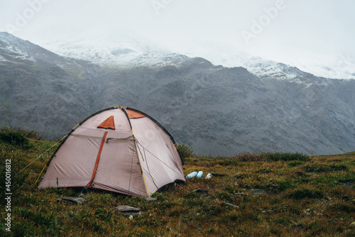 Scenic alpine landscape with tent on green hill among rocks. Beautiful snow-capped mountains in low clouds. Atmospheric scenery with tent in highlands in overcast weather. High mountains covered snow.