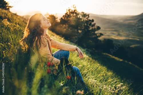 Young woman traveler with long loose curly hair sits on green grass meadow with flowers and types on smartphone against hilly landscape under sunlight photo
