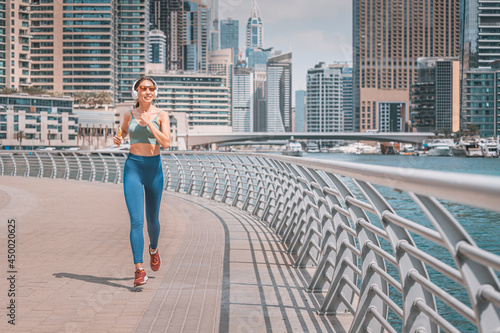 Happy woman wearing headphones and activewear top exercising and does running and fitness on the sidewalk of the embankment in the Dubai Marina area © EdNurg