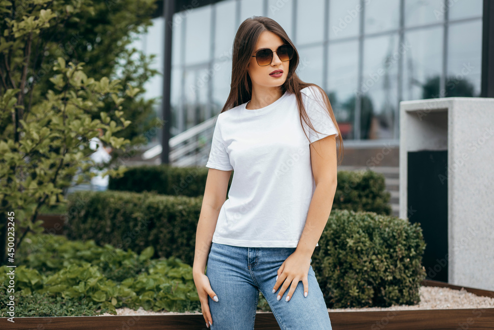 Fototapeta premium Stylish brunette girl wearing bwhite t-shirt and glasses posing against street , urban clothing style. Street photography