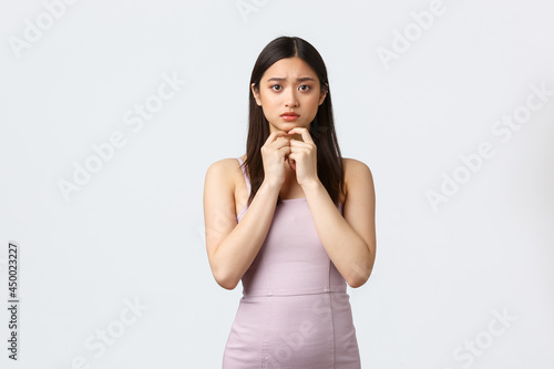 Celebration, beauty and glamour concept. Worried and cocnerned asian female in evening dress, elegant woman attent party, feeling uneasy and frustrated, looking nervous, white background photo