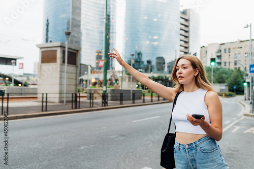 Young woman outdoors hailing taxi holding smartphone hitching