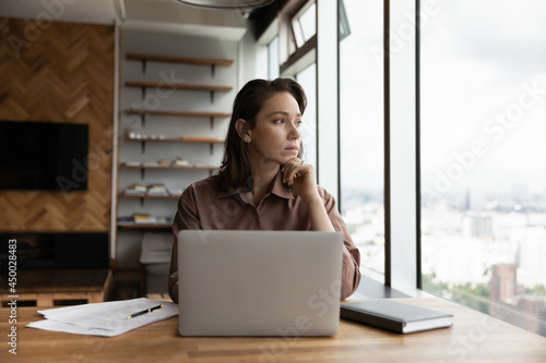 Thoughtful business owner woman looking out of window at city, sitting at workplace with laptop, thinking over solutions, challenges, company future vision, making decision, feeling uncertain