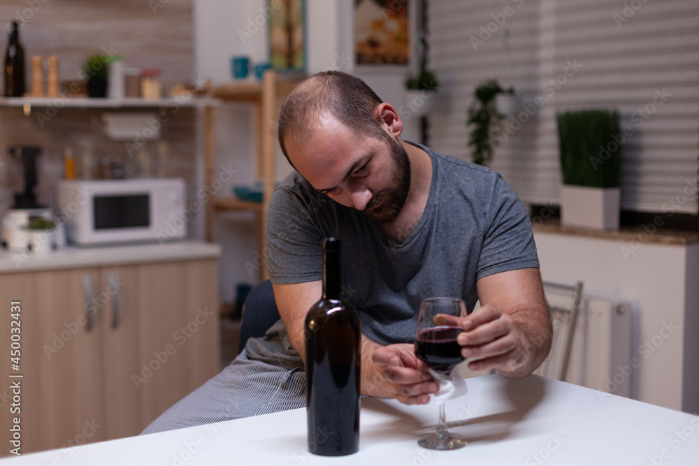 Caucasian man holding glass of wine sitting in kitchen at home. Depressed person drinking liquor, booze, alcoholic beverage alone while feeling intoxicated with bottle of alcohol Caucasian man holding glass of wine sitting in kitchen at home. Depressed person drinking liquor, booze, alcoholic beverage alone while feeling intoxicated with bottle of alcohol