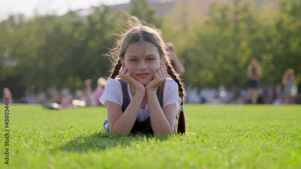 Smiling cute girl with braids lying on green lawn in park looking at camera Smiling cute girl with braids lying on green lawn in park looking at camera