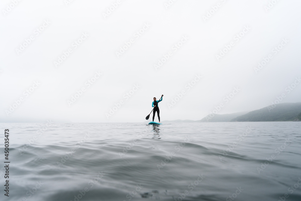 Fitness woman standing on surfboard enjoying supsurfing with paddle at endless sea water fog Fitness woman standing on surfboard enjoying supsurfing with paddle at endless sea water fog