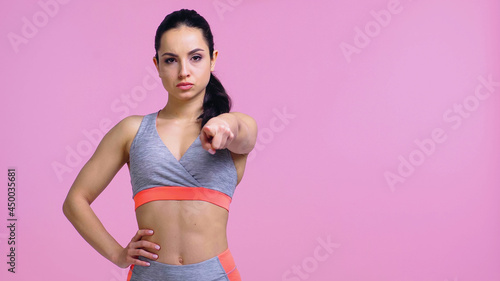 serious sportswoman standing with hand on hip and pointing with finger isolated on pink