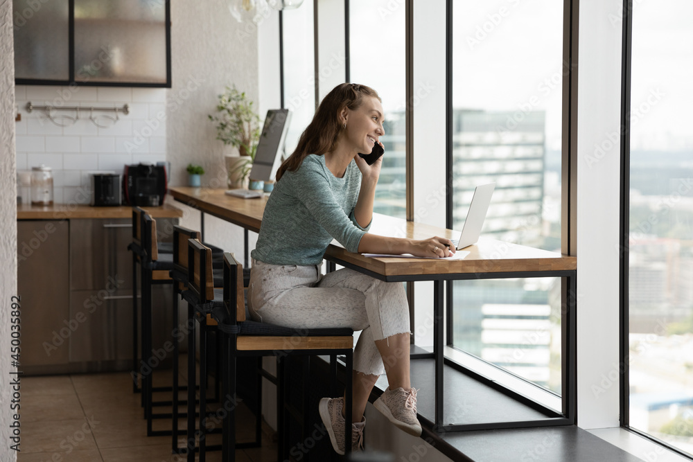 Happy millennial female manager, employee, entrepreneur working at laptop at city window, talking on mobile phone, giving answer, consultation to client. Gen Z student girl having nice phone call Happy millennial female manager, employee, entrepreneur working at laptop at city window, talking on mobile phone, giving answer, consultation to client. Gen Z student girl having nice phone call