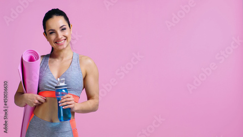 cheerful sportswoman holding fitness mat and sports bottle isolated on pink