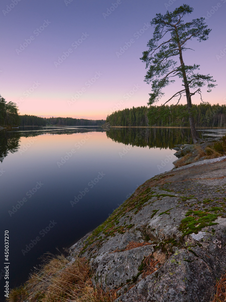 Obraz premium Granite stones, pine trees and forest are reflected in the clear serene waters of the Finnish lake.