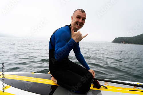 Portrait of smiling male surfer in wetsuit posing with paddleboarding board and paddle performing shaka gesture. © Aboltin