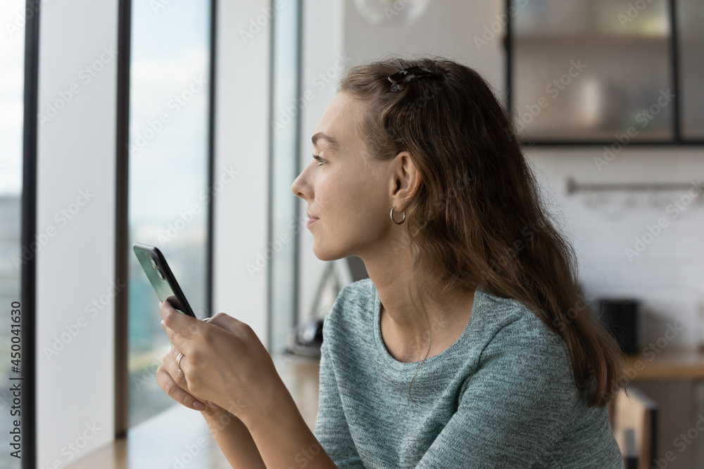 Thoughtful gen Z teen girl holding smartphone, looking out window in deep thoughts, thinking over text message, online chat talk, future meeting or dating, using online app on mobile phone Thoughtful gen Z teen girl holding smartphone, looking out window in deep thoughts, thinking over text message, online chat talk, future meeting or dating, using online app on mobile phone