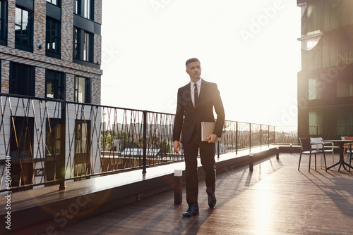 Confident mature man in full suit carrying laptop while walking by business center terrace outdoors