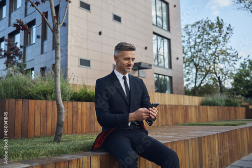 Confident mature businessman using smart phone while sitting outdoors near office building