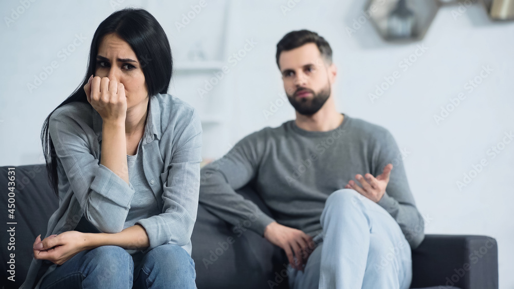 worried woman covering face and sitting near blurred and displeased boyfriend quarrelling in living room worried woman covering face and sitting near blurred and displeased boyfriend quarrelling in living room