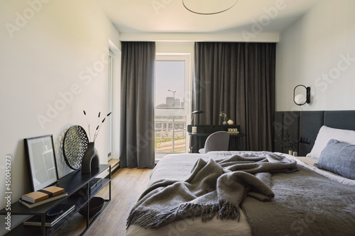 Stylish composition of small modern bedroom interior. Bed, creative lamp and elegant personal accessories. Walls with black panels. Panoramic windows. Minimalistic masculine concept. Template.