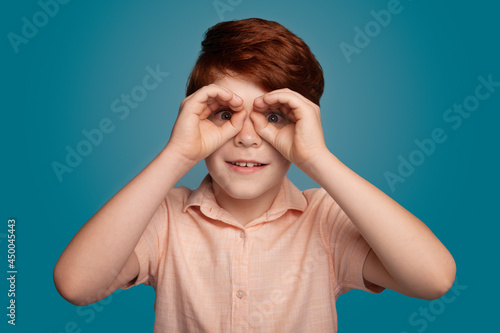 Ginger boy gesturing binocle sign on studio in trendy style. Fashion model.