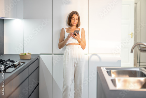 Young woman dressed in white standing relaxed with phone on the modern kitchen at home. Smart home and modern living concept