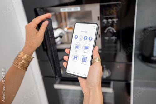 Smart phone with running mobile application to control smart devices in the kitchen. Female controlling microwave or oven with phone at home. Smart home concept