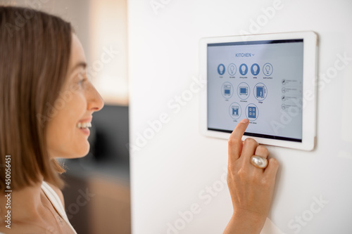 Happy woman controlling smart devices using control panel mounted on the wall at kitchen. Digital tablet with running mobile application. Smart home concept