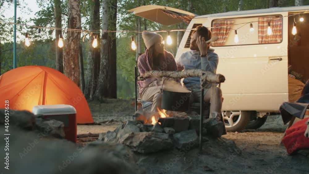 Young couple sitting at campsite in evening and speaking while cooking meal in pot over campfire Young couple sitting at campsite in evening and speaking while cooking meal in pot over campfire