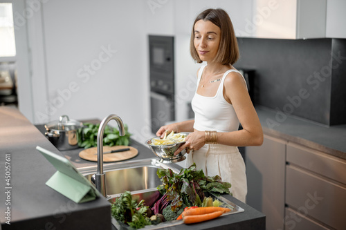 Young woman cooking with fresh vegetables and greens looking at recipe on the digital tablet at modern kitchen. Fresh organic ingredients, hygiene and healthy eating concept