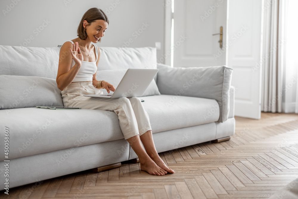Young woman talking online on laptop, having a video conference while sitting on the comfortable couch at white and cozy living room at home Young woman talking online on laptop, having a video conference while sitting on the comfortable couch at white and cozy living room at home