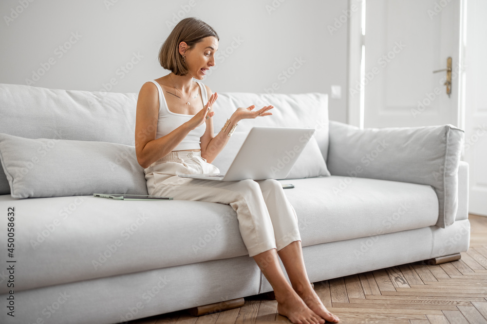 Young woman talking online on laptop, having a video conference while sitting on the comfortable couch at white and cozy living room at home Young woman talking online on laptop, having a video conference while sitting on the comfortable couch at white and cozy living room at home