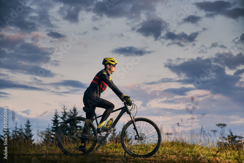 Young man cyclist riding bike on sunset in the mountains. Bicyclist wearing helmet, sports glasses and uniform. Side view. Beautiful evening sky on background. Concept of active lifestyle