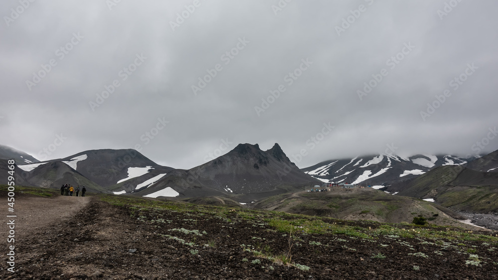 A dirt road runs through the valley. Ahead, against the background of a cloudy sky, you can see a bizarre mountain range. Snow on the slopes. A group of tourists goes to the mountains. Kamchatka