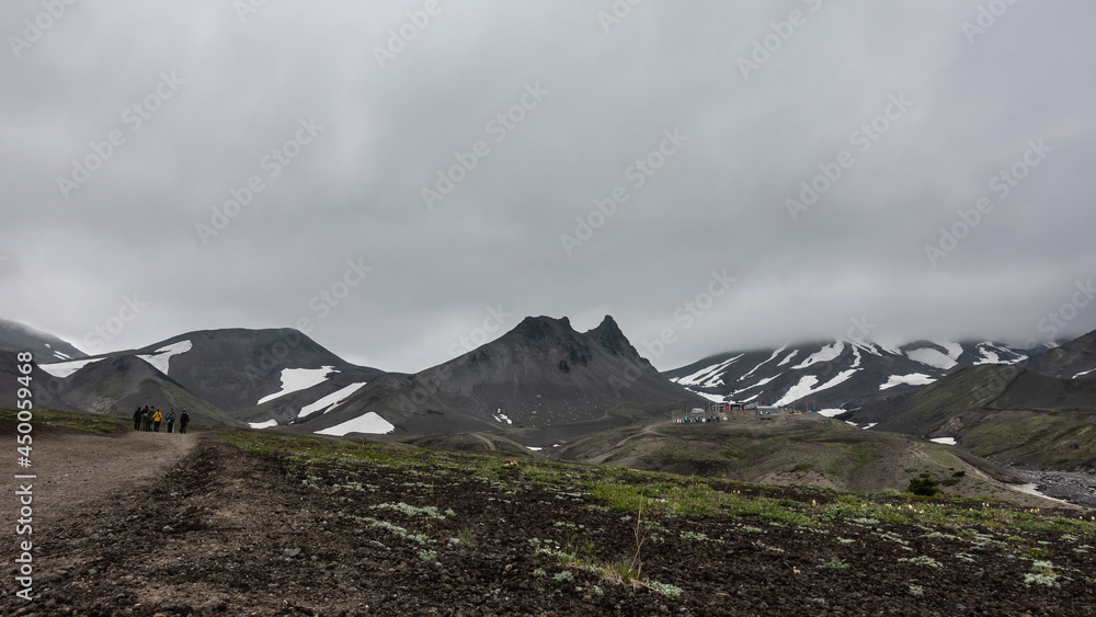 A dirt road runs through the valley. Ahead, against the background of a cloudy sky, you can see a bizarre mountain range. Snow on the slopes. A group of tourists goes to the mountains. Kamchatka A dirt road runs through the valley. Ahead, against the background of a cloudy sky, you can see a bizarre mountain range. Snow on the slopes. A group of tourists goes to the mountains. Kamchatka
