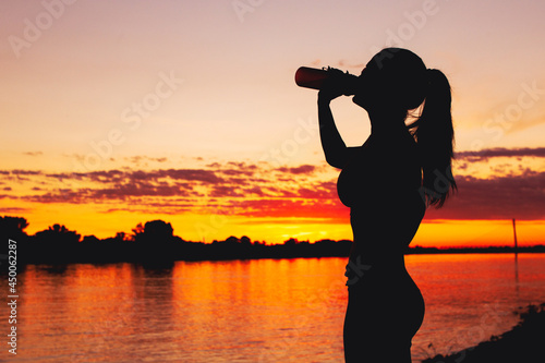 Thirsty woman silhouette drinking refreshment from bottle at riverbank in sunset