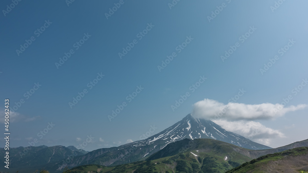 The conical stratovolcano Vilyuchinsky rises against the background of the blue sky. There is snow on the slopes. There is a fluffy white cloud over the top. A summer day. Kamchatka
