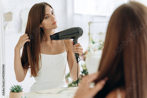 Beautiful girl using a hair dryer and smiling while looking at the mirror. Smiling woman drying hair with hair dry machine. Happy girl looking at mirror while using hair dryer in the bathroom