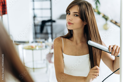 Quadro em tela Beautiful young smiling woman using a hair straightener while looking into the mirror in bathroom