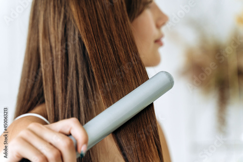 Hairdressing. Woman with beautiful long straight hair using hair straightener. Gorgeous girl straightening healthy hair with flat iron. Hair ironing and hairstyle concept