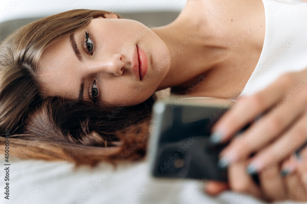 Obraz premium Romantic woman looking at mobile phone while laying on her bed. Brunette young woman using cellphone at home. Beautiful girl surfing the net on smartphone and relaxing