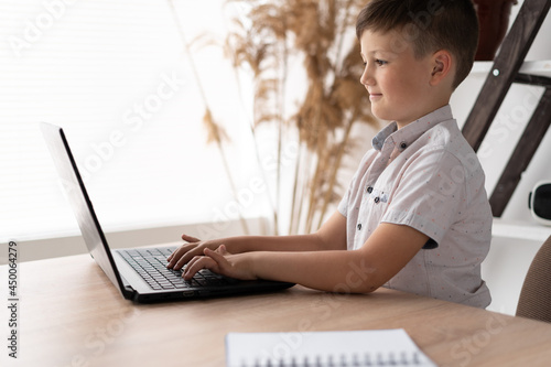 Side view of a schoolboy at home schooled working on a laptop typing text on the keyboard while communicating with a remote teacher. The child is studying computer programs online. photo