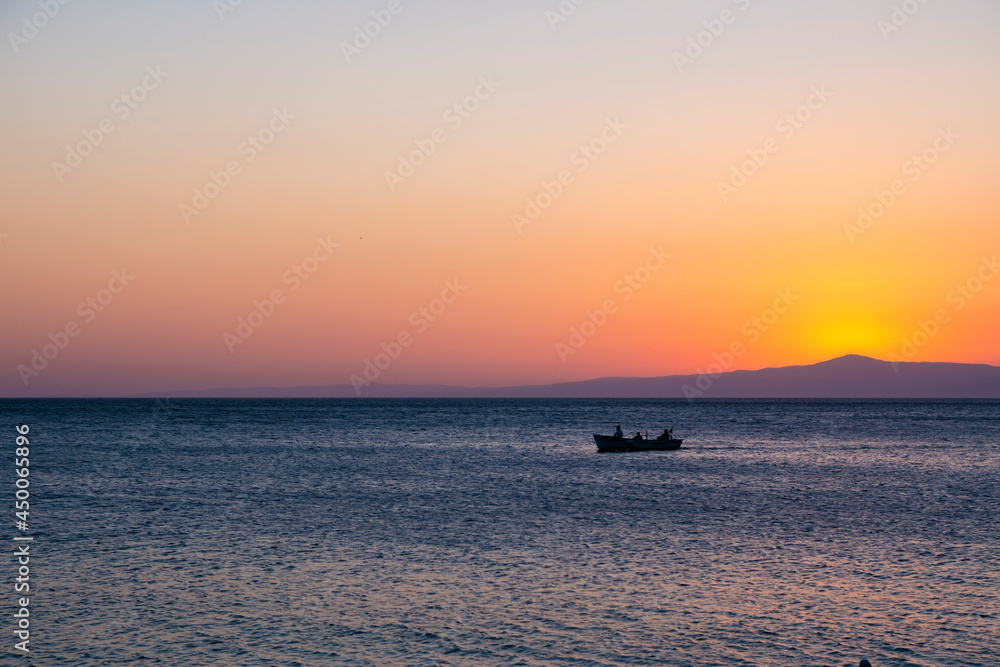 Fishermen in a tiny fisher boat with floating on peaceful blue Aegean Sea during sunrise, sunset. Dramatic orange sky and romantic island background with large copy space. Tranquility and serenity.