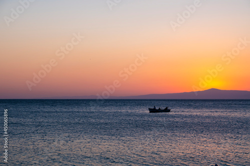 Fishermen in a tiny fisher boat with floating on peaceful blue Aegean Sea during sunrise, sunset. Dramatic orange sky and romantic island background with large copy space. Tranquility and serenity.