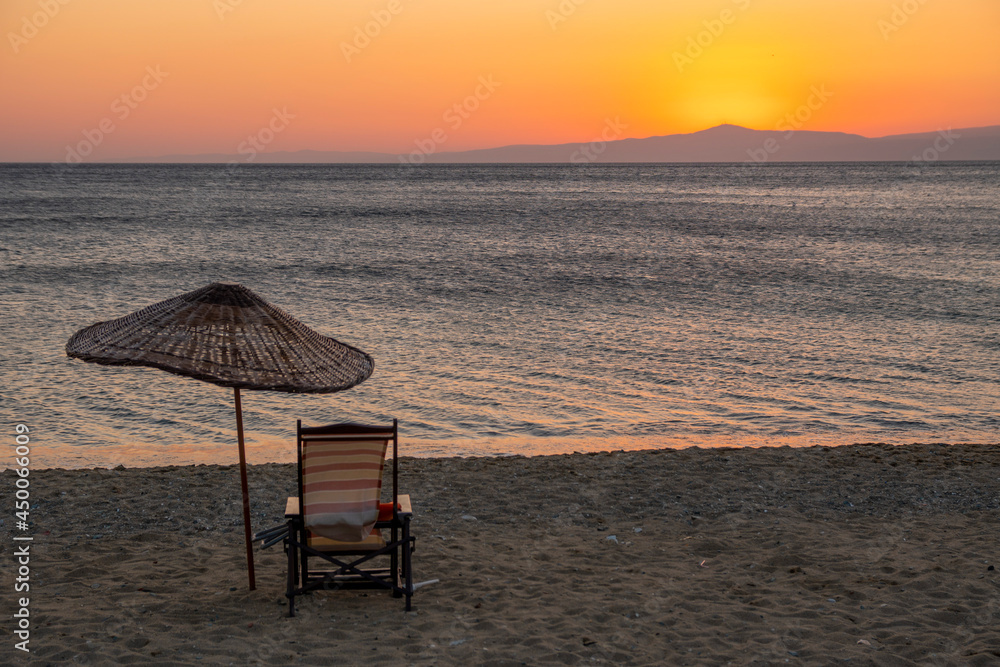 A single straw textured sunshade umbrella and an empty sun lounger standing on the coast at sunset after  everybody left home. Sand beach with orange sunset and peaceful sea background at dawn.