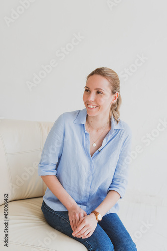 young blonde woman in a blue shirt sits on a sofa in a bright room and laughs