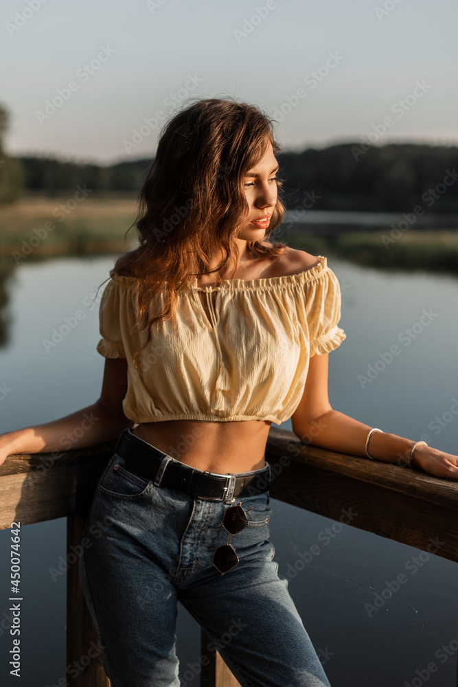 Fashionable beautiful woman model with curly hair in vintage blue jeans with summer fashionable blouse top stands on a wooden pier near a lake at sunset