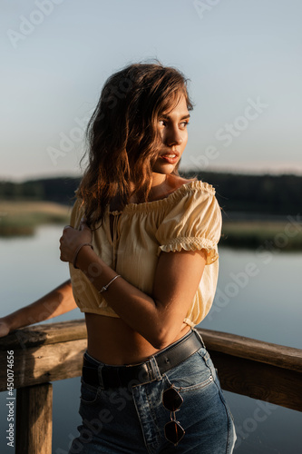 Nature fresh portrait of pretty young woman model with curly hair in blue jeans and yellow top blouse resting in nature near a lake at sunshine