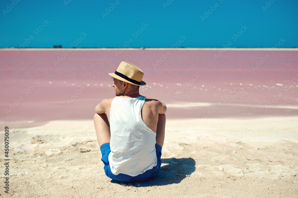 Back view of unrecognizable man, wearing white tshirt and straw hat, sitting against pink lake and clear blue sky, minimalism concept