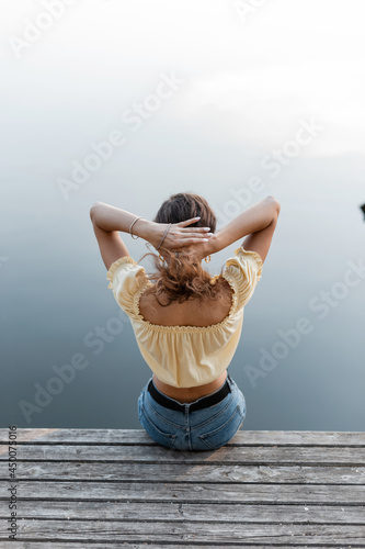 Young girl in fashionable denim clothes straightens her hair and sits on a wooden pier near the water photo