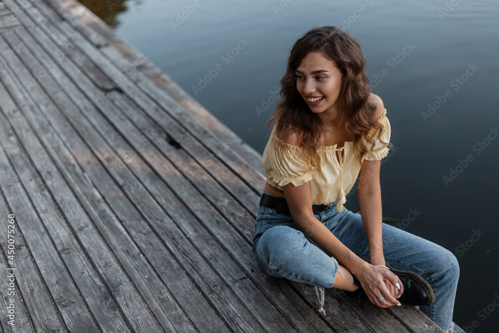 happy beautiful young girl with curly hairs in fashionable blue jeans and yellow blouse sits on a wooden pier